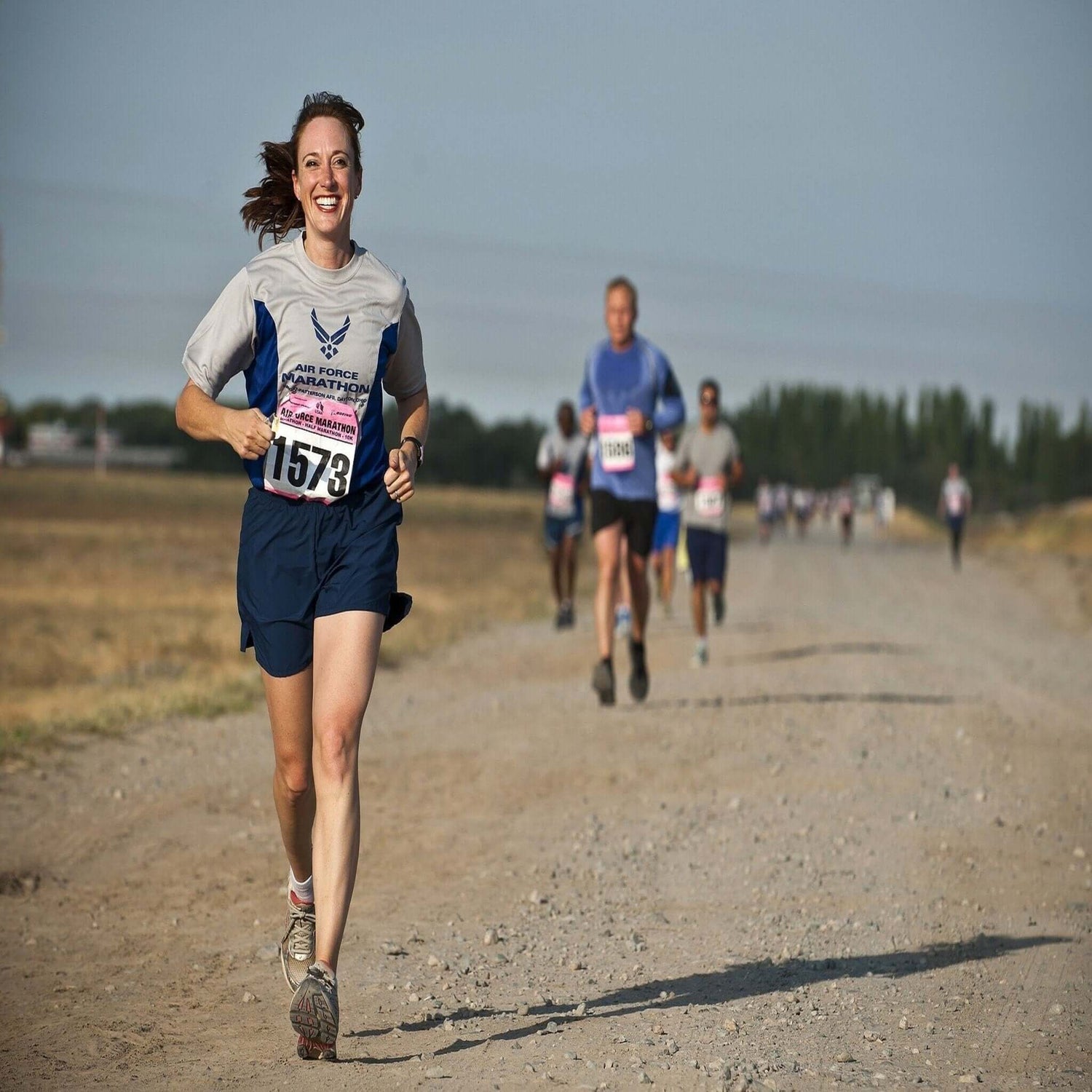 Picture of a woman running, leading the pack. Other runners are far behind her, on a long dusty track. She is smiling as she is winning the race.