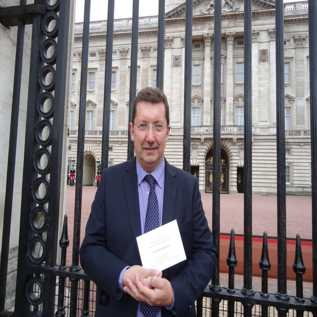 Photo of Michael Mahoney outside the gates of Buckingham Palace holding his invitation to tea with Queen Elizabeth II 